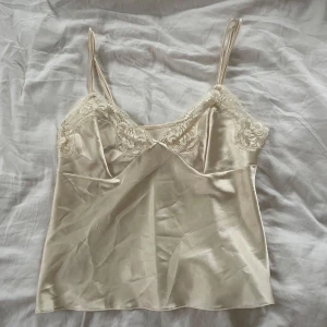 Beige satinlinne med spetsdetaljer - Elegant beige linne i satin med smala axelband och vacker spetsdetalj vid bysten. Perfekt för en stilren och feminin look.