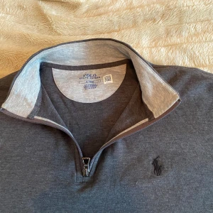 Grå tröja med dragkedja från Polo Ralph Lauren - Snygg grå tröja från Polo Ralph Lauren med halv dragkedja och broderad logga på bröstet. Perfekt för en stilren och avslappnad look. Tröjan har långa ärmar och en bekväm passform.