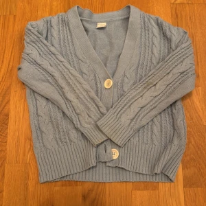 Ljusblå kofta från Lindex - Mysig ljusblå kofta från Lindex med kabelstickat mönster och stora knappar framtill. Perfekt för kyliga dagar med sin mjuka och bekväma passform. Passar barn i storlek 134/140. Eller 8-10 år gamla! I bra skick, men jag säljer eftersom den inte kommer till användning längre.