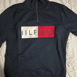 Mörkblå tröja med dragkedja från Tommy Hilfiger - Säljer en mörkblå tröja från Tommy Hilfiger med halv dragkedja och det ikoniska logotyptrycket i rött, vitt och blått på bröstet. Tröjan har en ribbad krage och är perfekt för en stilren look.