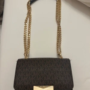 Brun axelväska från Michael Kors - Snygg brun axelväska från Michael Kors med guldfärgade kedjehandtag och logotypdetalj. Sista bilden visar repor som den har. 