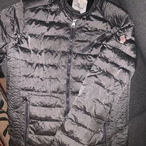 Grön dunjacka från Moncler - Säljer en stilren grön dunjacka från Moncler med dragkedja och långärmad design. Jackan har en quiltad yta och Moncler-logga på ärmen. Perfekt för kyligare väder.