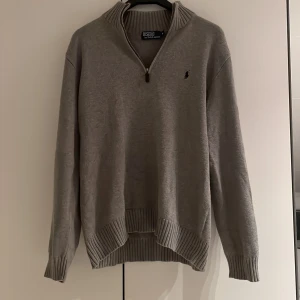 Grå tröja med dragkedja från Polo Ralph Lauren - Säljer en stilren grå tröja från Polo Ralph Lauren med halv dragkedja och ribbade detaljer vid krage, ärmslut och nederkant. Perfekt för en avslappnad men snygg look. 🐎