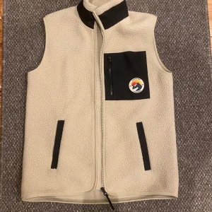 Beige fleeceväst från Everest - Säljer en beige fleeceväst från Everest med svart dragkedja och bröstficka. Västen har en broderad logga på bröstet och svarta detaljer vid fickorna. Perfekt för kyliga dagar.