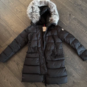 Moncler APHROTI HOODED SHEARLING jacka  - Snygg svart dunjacka från Moncler med en lyxig pälsdetalj på huvan. Jackan har dragkedja och knappar framtill samt praktiska fickor med dragkedjor. Perfekt för kalla vinterdagar. Storlek 00/XS nypris ca 23.000