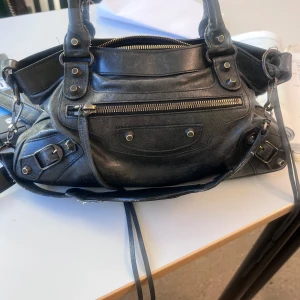 Balenciaga City Bag - Snygg svart handväska i skinn med flera dragkedjor och detaljer. Väskan har dubbla handtag och en avtagbar axelrem. Perfekt för att bära med sig det viktigaste. Den har en praktisk ficka med dragkedja på framsidan. Så fin o i jätte bra skick! Finns äkthetsbevis och kvitto, väskan är köpt på collectors cage där alla väskor är äkta! Pris går att diskutera🥰