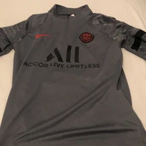 Grå psg tröja från nike  - Snygg grå fotbollströja från Nike med Paris Saint-Germain-logga på bröstet och 'Accor Live Limitless' tryckt på framsidan. Tröjan har korta ärmar med ett mönster och 'Visit Rwanda' tryckt på ryggen. Perfekt för PSG-fans!
