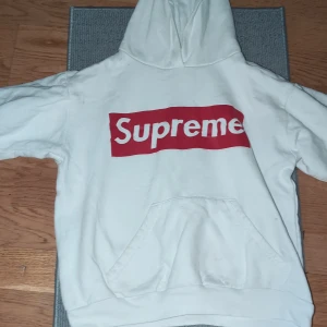Vit hoodie från Supreme - Säljer en vit hoodie från Supreme i storlek .134Den har en klassisk röd Supreme-logga på framsidan och en bekväm känguruficka. Perfekt för streetwear-stilen och att hålla sig varm under kyliga dagar. Passar både till vardags och chillhäng. 🧢