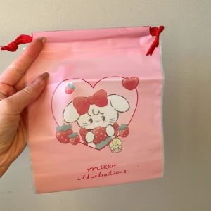 Rosa påse med illustration - Söt rosa påse med en illustration av en kanin med jordgubbar och en rosett. Påsen har röda snören för att dra åt öppningen. Perfekt för småsaker eller som presentpåse. Storlek 20x16 cm