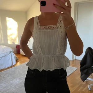 Vit ärmlös blus med spetsdetaljer - Säljer en söt vit blus med spetsdetaljer och volangkant. Blusen är ärmlös och har en luftig passform, perfekt för varma dagar. Den har ett diskret randigt mönster och en vacker spets runt halsringningen.