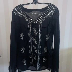 Svart blus med paljettdetaljer - Elegant svart blus med vackra silverpaljetter och broderade blommönster. Blusen har långa ärmar och en rund halsringning med dekorativa detaljer. Perfekt för en festlig kväll.