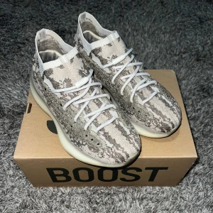 Yeezy Boost 380 - Säljer ett par Adidas Yeezy Boost 380 med ett unikt mönster i beige och grått. Skorna har en strumpliknande passform med snörning och en bekväm Boost-sula. Perfekta för dig som vill ha både stil och komfort.
