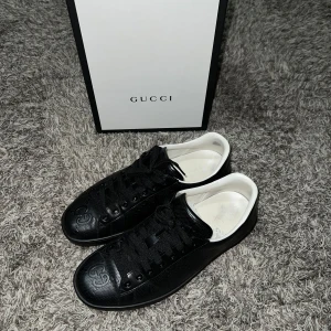  Gucci sneakers  - Snygga svarta sneakers från Gucci med diskret logotyp på sidan och tå. Skorna har en klassisk design med snörning och vit insida. Perfekta för en stilren look.