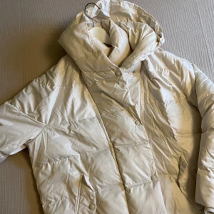 Vit pufferjacka från Zara - Säljer en stilren benvit pufferjacka från Zara. Jackan har en hög krage och dragkedja framtill. Den är långärmad och har en quiltad design. 