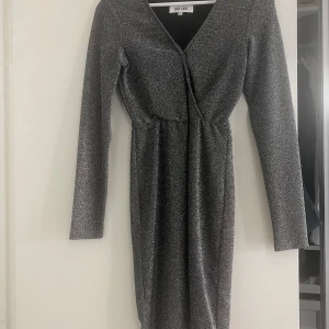 Glittrig omlottklänning - Snygg glittrig klänning med omlottdesign och v-ringning. Klänningen har långa ärmar och en figurnära passform som framhäver silhuetten. Perfekt för en kväll ute!