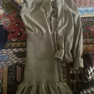 Beige långklänning med volangdetaljer - Elegant beige långklänning med smockad midja och volangdetaljer vid fållen. Klänningen har långa ärmar med elastiska ärmslut för en bekväm passform.