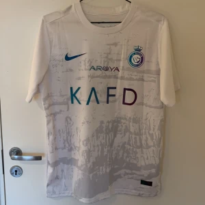 Al nassr tröja Cristiano Ronaldo - Säljer en vit fotbollströja från Nike med Al Nassr FC-logga och KAFD-tryck på framsidan. Har Ronaldo på ryggen. Tröjan är deras tredje kit. Storlek: M. Skick: 10/10