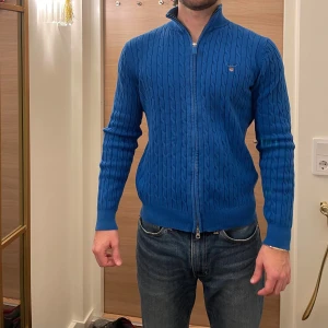 Blå stickad tröja med dragkedja från GANT - Snygg blå stickad tröja från GANT med dragkedja och kabelstickat mönster. Perfekt för kyligare dagar med sin stilrena design och bekväma passform.