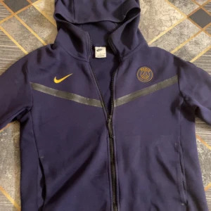 Mörkblå zip-hoodie från Nike med PSG-logga - Säljer en mörkblå hoodie från Nike med Paris Saint-Germain-logga. Hoodien har dragkedja och huva, samt en gul Nike-swoosh på bröstet. Perfekt för fotbollsfans! OBS den har defekt på grund av tvätt kolla bild 5. Priset är inte hugget i sten.