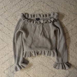 Grå tröja från Bubbleroom  - Säljer en stilren grå stickad tröja från Bubbleroom med volangdetaljer vid hals, ärmslut och nederkant. Tröjan har långa ärmar och en offshoulder-design som ger en trendig look. Supersnygg och perfekt för kyligare dagar!