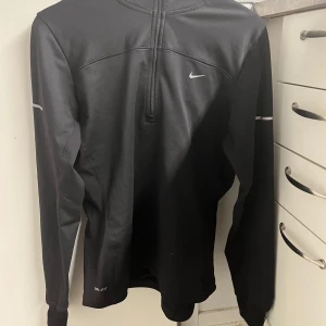 Svart långärmad tröja från Nike - Snygg svart långärmad tröja från Nike med dragkedja och reflekterande detaljer. Perfekt för träning med bekväm passform och tumhål vid ärmsluten. Nike-logga i reflex