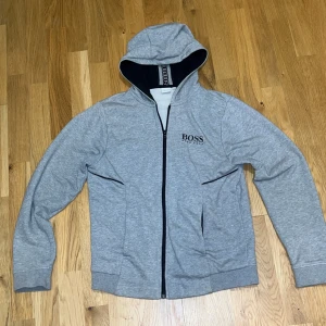 Grå hoodie från Boss - Säljer en snygg grå hoodie från Boss i storlek M(Skulle absolut säga att den är S). Den har en dragkedja framtill och en bekväm huva. Perfekt för en avslappnad stil och passar bra till både höst och vår. Boss-loggan är diskret placerad på bröstet.