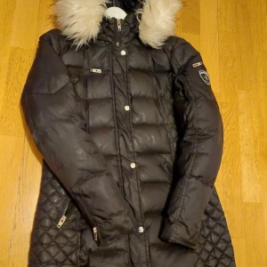 Svart dunjacka med pälsdetaljer - Säljer en snygg svart dunjacka från rockandblue, storlek dam L, i mycket bra skick. Jackan har en fluffig pälsdetalj på huvan och quiltade mönster på sidorna. Den är perfekt för vintern med sina långa ärmar och dragkedja framtill. Den enda synliga deffekten är en liten hål på ena armen som man kan se på bild 4. Dock syns den inte väldigt mycket i verkligheten. 