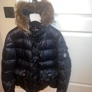 Säljer en snygg svart dunjacka från Moncler i mycket bra skick. Jackan har en avtagbar huva med pälsdetaljer och praktiska fickor med knappar. Perfekt för kalla vinterdagar med sin quiltade design och dragkedja framtill. Passar både till vardags och festliga tillfällen. Dessvärre behöver dragkedjan att bytas ut 