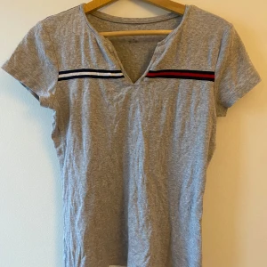 Grå t-shirt med ränder - Säljer en grå t-shirt från Tommy Hilfiger i bra skick. Den har en v-ringning och korta ärmar. Två ränder i svart och rött pryder bröstet