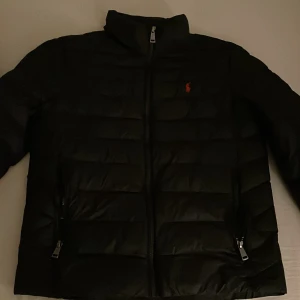 Svart pufferjacka från Ralph Lauren - Säljer en snygg svart pufferjacka från Ralph Lauren. Jackan har en dragkedja framtill och är quiltad för extra värme. Perfekt för kyliga höst- och vinterdagar. Den har en klassisk design med en liten logga på bröstet. Passar både till vardags och lite finare tillfällen.
