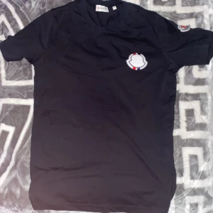 Svart t-shirt från Moncler - Säljer en snygg svart t-shirt från Moncler i storlek S. Den har ett coolt Moncler-märke på bröstet och ärmen. Perfekt för en stilren och avslappnad look. Passar bra till både vardag och fest!