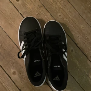 Svarta Adidas sneakers - Säljer ett par snygga svarta Adidas sneakers i mycket bra skick. De har de klassiska tre vita ränderna på sidan och vit sula. Perfekta för vardagsbruk och passar till nästan alla outfits. Skorna har snörning och är bekväma att gå i. Perfekta för både vår och höst!