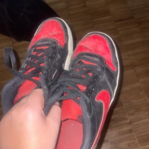 Svarta och röda Nike sneakers - Säljer ett par svarta och röda Nike sneakers i bra skick. De har en klassisk design med svart ovandel och röda detaljer, inklusive den ikoniska swoosh-loggan. Skorna har svarta snören och en röd sula. Perfekta för vardagsbruk eller träning!