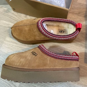 UGG TAZZ  - Säljer ett par snygga bruna UGG boots i mocka med en röd och vit kantdetalj. De är i nytt skick och kommer i originalkartong.💌Storlek: 39