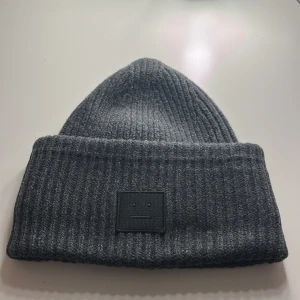 Grå ribbad mössa från Acne Studios - Säljer en snygg grå ribbad mössa från Acne Studios som gått ut ur sortimentet. Den är i ullblandning med 97% ull, vilket gör den perfekt för kalla dagar. Mössan har en stilren design med en diskret logga framtill. Perfekt för både höst och vinter!