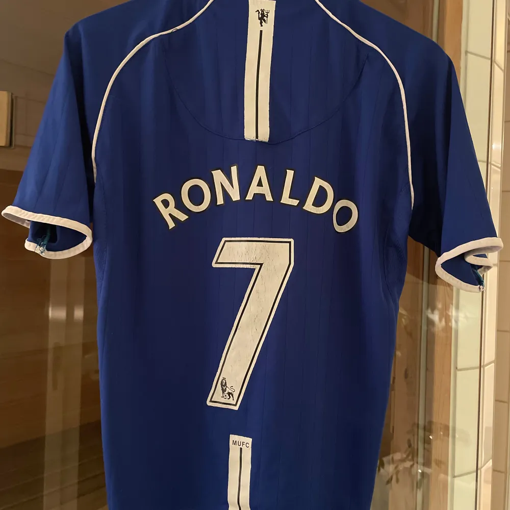 Säljer en blå Manchester United fotbollströja med korta ärmar och vit kant. Den har ett tryck av 'Ronaldo' och nummer 7 på baksidan. Perfekt för fans av laget och spelaren! Tröjan är i bra skick.. T-shirts.