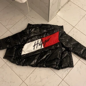 Svart pufferjacka från Tommy Hilfiger - Säljer en snygg svart pufferjacka från Tommy Hilfiger. Jackan har en glansig finish och ett stort Hilfiger-tryck i rött och vitt på ryggen. Den är perfekt för kalla dagar och har en dragkedja framtill. Jackan är i mycket bra skick och redo för vintern!