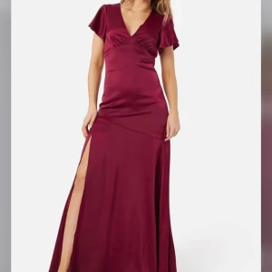 Vinröd långklänning med slits - Elegant vinröd långklänning i satin med en djärv slits på sidan. Klänningen har kortärmade volanger och en v-ringad design som ger en sofistikerad look. Perfekt för festliga tillfällen så som bal. Prislappen sitter kvar 💕