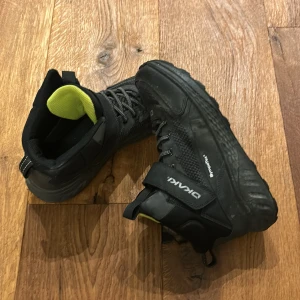 Svarta sneakers från DKNY - Säljer ett par svarta sneakers från DKNY i bra skick. De har en cool design med snörning och en extra rem för bättre passform. Skorna har en robust sula och är perfekta för höst och vinter. Insidan är fodrad med en grön detalj som ger en snygg kontrast.