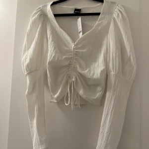 Vit långärmad topp från Gina Tricot - Säljer en superfin vit långärmad topp från Gina Tricot i storlek S. Toppen har en snygg rynkad detalj framtill med knytning och är i ett mjukt, ribbat material. Perfekt för både vardag och fest! ✨
