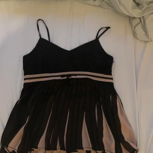 Svart och beige klänning med axelband - Säljer en elegant svart topp med tunna axelband och beige detaljer. Den har en vacker omlott-design med en lätt plisserad kjol. 