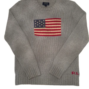 Grå stickad tröja från Polo Ralph Lauren - Säljer en snygg grå stickad tröja från Polo Ralph Lauren med en amerikansk flagga på bröstet. Tröjan har långa ärmar och RL-broderi i rött vid nederkanten. Perfekt för höst och vinter!