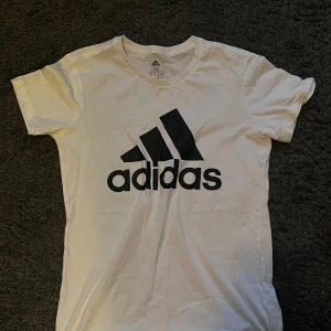 Vit t-shirt från Adidas - Säljer en snygg vit t-shirt från Adidas i storlek XS. Den har en stor svart Adidas-logga på framsidan. Passar både till vardags och sportiga aktiviteter. Använd ett fåtal gånger men inget som syns.