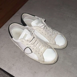 Vita sneakers från Philippe Model - Säljer ett par vita sneakers från Philippe Model i bra skick. De har svarta detaljer på hälen och klassisk snörning. Perfekta för vardagsbruk och stilrena till alla outfits. Sulan visar lite användning men är fortfarande i gott skick. 1999 först till kvarn