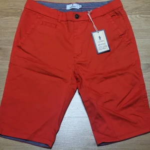 Röda shorts från Chisnbal - Snygga röda shorts från Chisnbal i bomull. De har en knapp och dragkedja framtill samt praktiska fickor. Perfekta för sommardagar och avslappnade tillfällen. Broderad detalj på baksidan ger en extra touch.