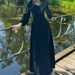 Svart långklänning med slits - Elegant svart långklänning med en stilren design och en smickrande slits på sidan. Klänningen har långa ärmar och en v-ringad halslinje som ger en sofistikerad look. Perfekt för höst- och vinterfester eller en stilfull kväll ute.