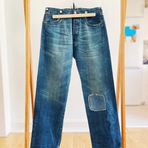 Double Back-Pocked Levis Japan - Deadstock 2003/4 Reprod Levis Vintage 501 Selvedge fr. 1918 - Tillverkad i USA 2003/4 hos Taylor Togs (fabrikskod '643' på Tag, förmodligen på baksidan av knappen), North Carolina som tog över produktionen från Levi's San Fransisco, Valencia Factory. De är från 1918 med en "Every Garment Guaranteed"-märke.   Midja: 82 cm (platt x 2) Innersöm: 78 cm Höjd: 33 cm Fållbredd: 25 cm Total längd: 107 cm Grennit - Nakna nitar - Hängselknapp - Solidgraverat vanligt spänne baktill med nål - Utsömmen vit söm - Kedjesöm