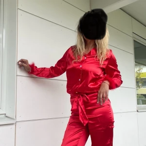 Röd satin pyjamas med fjäderdetaljer - Säljer en supercool röd pyjamas i satin med knappar och knytband i midjan. Den har unika fjäderdetaljer vid ärmslut och benslut som ger en extra touch. Perfekt för en festlig kväll hemma eller nyår! 🥳