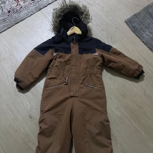 Brun overall med pälsfodrad huva från HM - Säljer en brun overall med svart överdel och pälsfodrad huva. Perfekt för kalla vinterdagar! Overallen har långa ärmar och dragkedja framtill. Den är i mycket bra skick och redo för nya äventyr i snön.