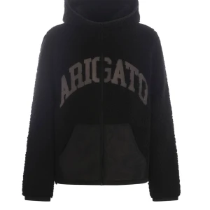 Svart teddyjacka Axel Arigato - Mysig svart teddyjacka med stor text 'ARIGATO' på framsidan. Jackan har en dragkedja och en stor huva för extra värme. Perfekt för kyliga dagar när du vill vara både bekväm och stilren. Den har en avslappnad passform och är supermjuk!